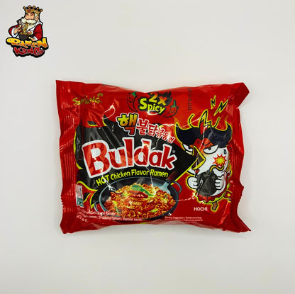 Ein ungeöffnetes Paket von Buldak 2x Spicy Ramen. Die Verpackung ist hauptsächlich rot mit einem Bild eines cartoonisierten Huhns, das Feuer spuckt, und mehreren koreanischen Schriftzeichen, die auf den scharfen Geschmack hinweisen. Die Packung ist umgeben von zahlreichen frischen roten Chilischoten.