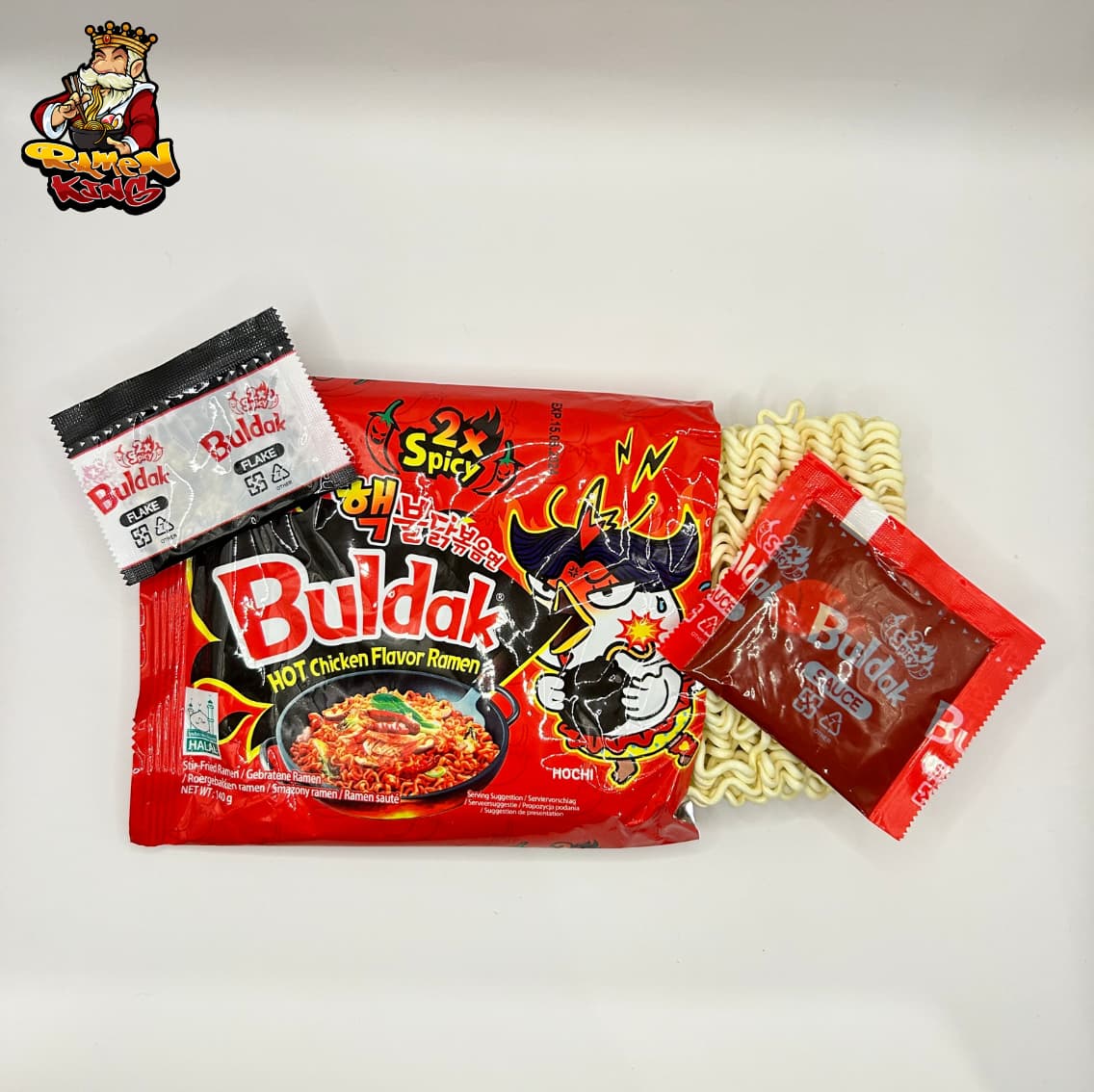 Geöffnetes Paket von Buldak 2x Spicy Ramen mit sichtbarem Inhalt. Zu sehen sind die Nudeln, ein schwarzer Beutel mit "Flake" und ein roter Beutel mit "Sauce", die alle neben der auffälligen roten Verpackung liegen.