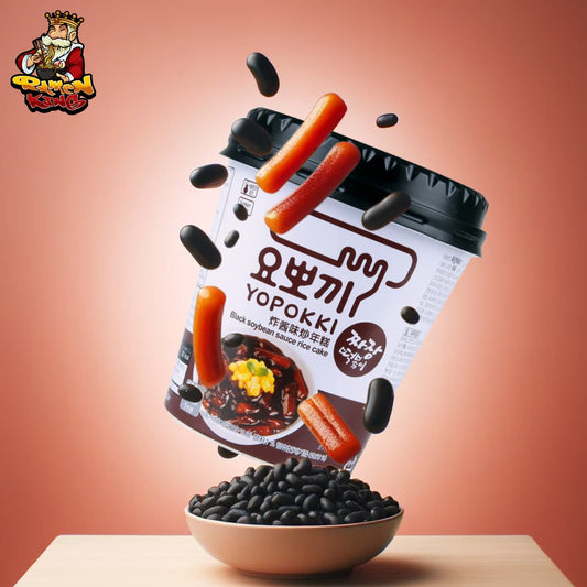 Ein Werbebild für Yopokki Jjajang Reiskuchen, bei dem eine Schüssel mit schwarzen Bohnen im Vordergrund steht und darüber ein umgestürzter Becher von Yopokki schwebt, aus dem die roten Reiskuchen und schwarze Bohnen in die Schüssel zu fallen scheinen. Der Becher hat die Aufschrift 'Yopokki' und darunter auf Koreanisch sowie 'Black soybean sauce rice cake'. Im Hintergrund ist eine neutrale, farblich abgestimmte Kulisse zu sehen, die das Produkt hervorhebt.